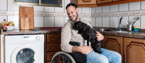 A man in a wheelchair is in his kitchen with a black dog on his lap.
